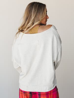 Shot from the back, she's wearing an oversized off-shoulder sweatshirt in heathered oatmeal with a worn-in, intentionally distressed hem — layered over a gold lace bralette and paired with a bright plaid skirt peeking out at the bottom.