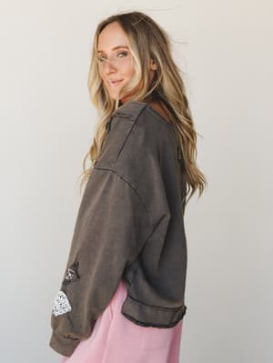 A model shot from a side-back angle showing an oversized acid-wash sweatshirt in olive charcoal with patches and lace trim details on the sleeve, paired with soft pink pants — casual and expressive with a vintage, lived-in feel.