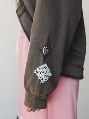 A close-up detail shot of a washed olive charcoal sweatshirt sleeve, layered over a soft pink skirt — the sleeve features a raw-edge lace patch and a small peace sign patch that give it that perfectly worn-in, personal feel.