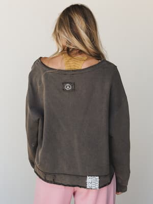 A back-view shot of a washed charcoal sweatshirt with raw-edge seams, a peace sign patch at center back, and a lace trim detail at the hem — layered over a mustard lace bralette and blush pants for that effortlessly collected, lived-in look.