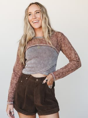 A layered long-sleeve floral lace top styled over a washed ribbed crop, paired with dark chocolate corduroy shorts and stacked silver rings — the kind of effortless mix-and-match that looks intentional without trying too hard.