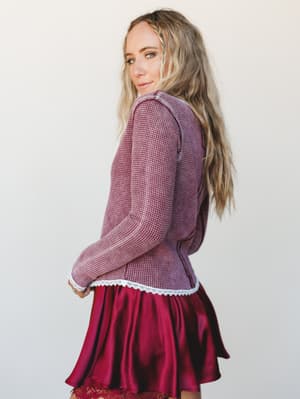 Shot from behind with a glance over the shoulder, she's wearing a mauve-red waffle knit top with white lace trim at the hem layered over a satin mini skirt in deep cranberry — the kind of mix-and-match that looks effortless but is clearly intentional