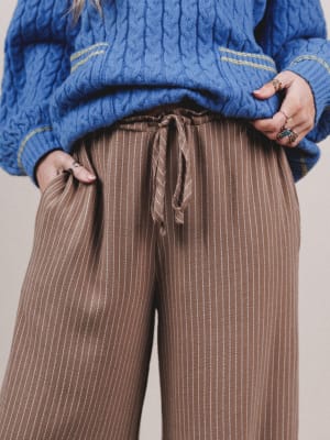 Person wearing a blue cable knit sweater and brown striped pants against a neutral background