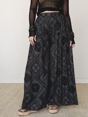 A moody, editorial-leaning mid-shot of wide-leg pants in a dark bandana-inspired paisley print, paired with a sheer polka-dot crop top and woven platform sandals — the kind of outfit that feels effortlessly free-spirited without trying too hard.