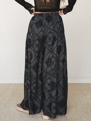 Shot from behind, a model wears voluminous wide-leg pants in a black paisley bandana print paired with a sheer black lace crop top and espadrille wedges — the whole look has that effortlessly moody, boho-after-dark energy.
