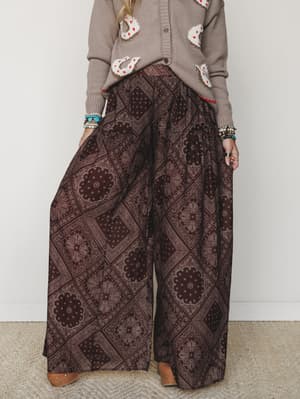 Wide-leg printed pants in a deep brown bandana-inspired print styled with a cozy graphic sweater and stacked bracelets — the kind of outfit that looks effortless but you definitely thought about it.
