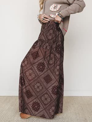 A wide-leg maxi pant in deep brown paisley-bandana print styled with a taupe graphic knit sweater and stacked turquoise and beaded bracelets — earthy, expressive, and very much the kind of outfit that makes people ask where you got that skirt.
