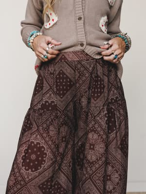 A close mid-shot of a stacked-jewelry moment — chunky silver rings, turquoise beaded bracelets, and a bandana-print wide-leg pant paired with a cozy intarsia cardigan, all in warm earthy tones against a soft neutral studio backdrop.
