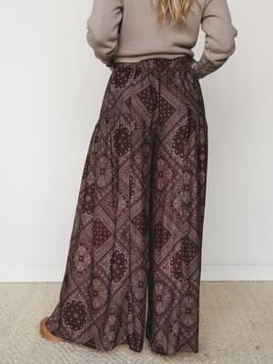 Shot from behind, a model wears sweeping wide-leg pants in a rich brown paisley-bandana print, paired with a taupe ribbed sweater — the kind of outfit that feels vintage-collected and genuinely comfortable at the same time.