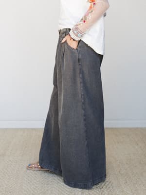 A side-profile shot highlighting wide-leg acid-washed denim trousers in charcoal black, styled with a white top featuring sheer floral-embroidered sleeves and layered bracelets — relaxed and expressive, the kind of outfit that looks effortless but wa
