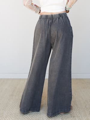 A back-view shot of wide-leg acid wash trousers in faded charcoal black, styled with a cropped white mesh-sleeve top and layered bracelets — relaxed and easy with just enough personality.