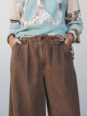 A mid-shot of wide-leg brown trousers styled with a layered waffle-knit top featuring a patchwork peace sign graphic, hands in pockets and stacked bracelets adding a relaxed, lived-in boho energy.