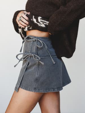 A close-up torso shot showing a washed grey denim mini skort with front tie details, paired with a cropped dark brown fair isle knit sweater and silver stacked rings — a lived-in, effortlessly cool combo that feels equal parts vintage and now.