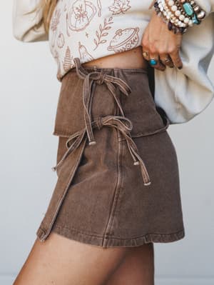 A close-up styled shot of a washed brown denim skort with lace-up tie details layered under a cream graphic sweatshirt, accessorized with stacked turquoise and pearl bracelets and rings — casual boho done right.