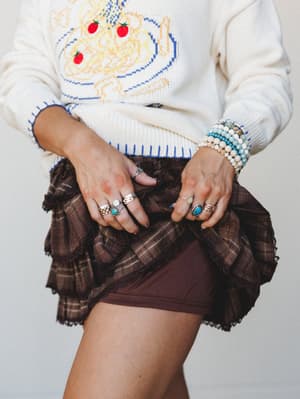 A close-up torso shot of a layered look — a cream graphic knit sweater with a playful embroidered bowl motif tucked over a ruffled brown plaid mini skirt, styled with stacked beaded bracelets and silver and turquoise rings for that lived-in, collecte