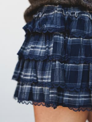 A close-up detail shot of a navy plaid mini skirt with three ruffled tiers, each finished with scalloped lace trim — the kind of piece that looks equally at home tucked into boots for a fall concert or layered over tights for a cozy night out.
