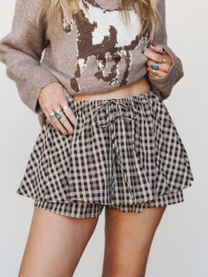 A close-up mid-shot of a gingham skort paired with a cow-print cropped sweater, styled with layered silver and turquoise rings — casual and collected with a little Western personality.