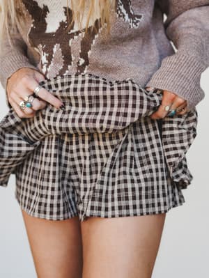 A close-up torso shot of a layered boho look — a plaid skort paired with a cow-print knit sweater, hands gathering the skirt fabric and stacked with silver and turquoise rings.