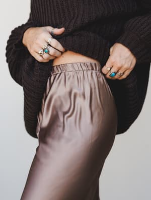 A close-up torso shot of a satin bronze trouser paired with a chunky dark brown cable-knit sweater, hands resting at the waistband and stacked with silver and turquoise rings — the kind of effortless mix-and-match that feels intentional without tryin
