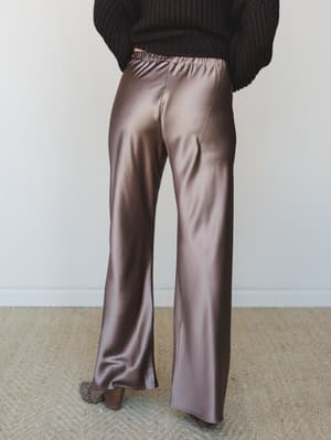 Shot from behind, a model wears wide-leg satin trousers in a warm bronze-mauve paired with a chunky dark knit sweater — the kind of outfit that looks equally at home at a dinner reservation or a lazy Saturday that got a little dressed up.
