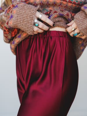 A close-up detail shot of a deep mulberry satin trouser paired with a chunky fair isle knit sweater, hands adorned with layered silver and turquoise rings pulling up the waistband — the kind of outfit that feels both intentional and effortless.