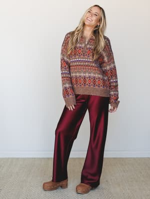 A laughing model pairs a cozy Fair Isle half-zip sweater with wide-leg satin trousers in deep mulberry — the kind of outfit that somehow works for both a holiday dinner and a Sunday you want to feel good about.