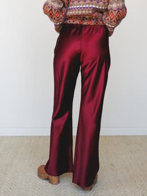 Shot from behind, a model wears fluid satin wide-leg trousers in a deep mulberry paired with a cozy fair isle sweater and wooden clogs — the kind of outfit that makes a Saturday feel intentional.