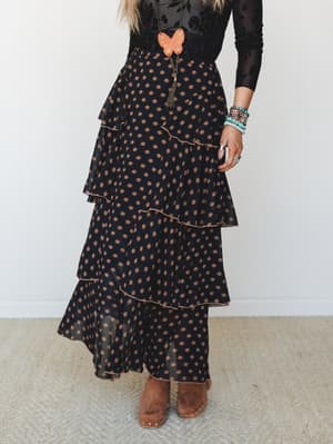 A tiered maxi skirt in black with warm rust daisy print styled with a sheer floral long-sleeve top, ankle boots, and stacked boho jewelry — the kind of outfit that looks equally at home at a fall market or a candlelit dinner.