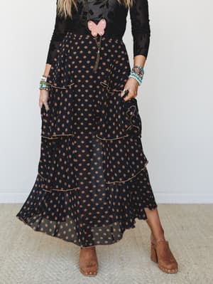 A tiered black maxi skirt with a warm bronze floral print styled with a sheer flocked top, layered turquoise bracelets, a pink butterfly pendant necklace, and tan block-heel clogs — boho-romantic with just enough edge.