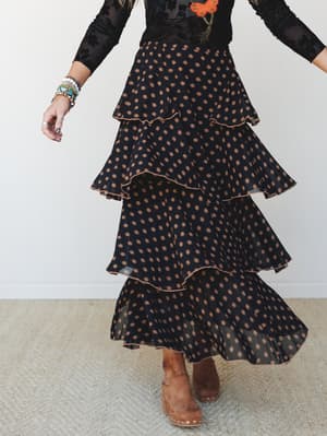 A tiered maxi skirt in black with warm tan floral dot print swirls mid-motion, paired with a sheer black floral top and tan suede clogs — the kind of outfit that looks equally at home at a fall farmers market or a night out with good people.