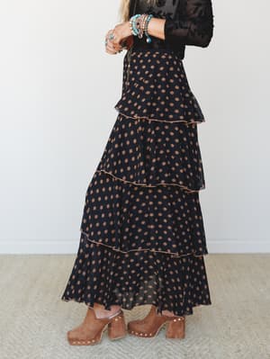 A tiered black maxi skirt with a warm-toned floral dot print styled with a sheer floral top, stacked turquoise bracelets, and studded suede clogs — boho layering done with intention.