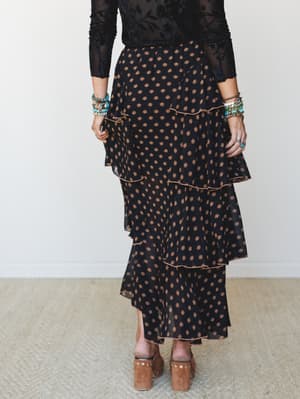 A tiered maxi skirt in black with warm tan floral medallion print, styled with a sheer black floral long-sleeve top, stacked turquoise and beaded bracelets, and tan suede platform clogs — the kind of outfit that feels effortless but clearly wasn't an