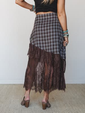 Shot from behind, a model pairs a brown plaid midi skirt with a sheer lace hem against a clean studio backdrop — the mix of flannel plaid and delicate lace trim gives it that effortless boho-romantic tension, finished with stacked turquoise bracelets