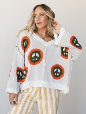An oversized cream knit sweater with chunky peace sign embroidery in rust orange and olive green, styled with yellow-and-white striped wide-leg pants — the kind of outfit that feels like a mood board for a really good decade.