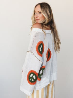 A breezy, lived-in knit sweater with oversized peace sign intarsia in rust and olive steals the show here — worn slightly off-shoulder with a side glance that feels effortlessly retro and just the right amount of carefree.