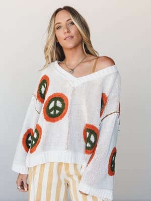A relaxed, oversized cream knit sweater covered in chunky peace sign patches in rust orange and olive green, worn off-the-shoulder over a tan cami and paired with yellow striped wide-leg pants — effortlessly retro and full of personality.