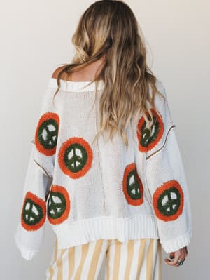 Shot from behind, this oversized cream knit sweater makes a statement with bold tufted peace sign patches in rust orange and olive green — worn off-shoulder over wide-leg butter yellow stripe pants, the whole look feels like a '70s afternoon done rig