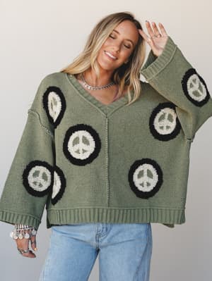 An oversized sage green knit sweater covered in tufted peace sign patches gets styled with light wash jeans and layered boho jewelry — the whole look has that effortless, 'grabbed it off the chair' energy that somehow works perfectly.