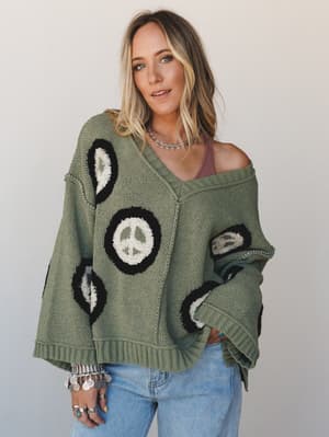 An oversized sage green knit sweater with chunky peace sign intarsia worn off-shoulder over a dusty pink cami, styled with light wash jeans and layered silver boho jewelry — the kind of sweater that does all the talking.