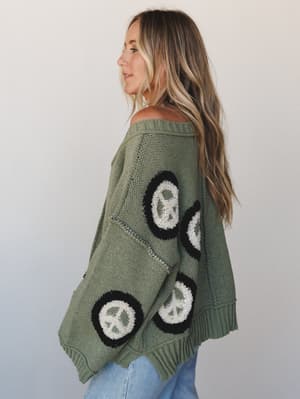 A relaxed, oversized knit sweater in muted olive green covered in chunky peace sign patches — worn slightly off-shoulder with jeans for that effortless, lived-in boho feel.