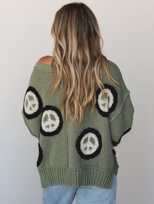 Shot from the back, a relaxed oversized knit sweater in muted olive green covered in chunky peace sign patches — the kind of piece that feels like it has a personality before you even put it on.