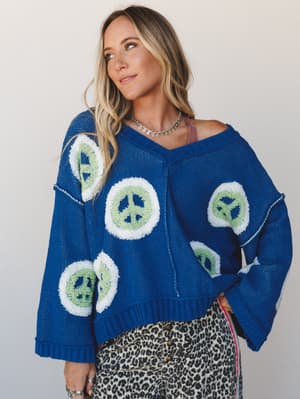 A cobalt blue oversized knit sweater with bold peace sign intarsia detailing, styled over a leopard print skirt with a delicate chain choker — the kind of outfit that has a point of view without trying too hard.