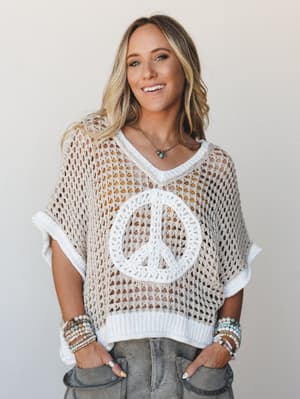 An open-knit crochet poncho top in cream with a bold peace sign graphic on the front, styled over a mustard layer with grey cargo pants and stacked boho bracelets — effortless, free-spirited, and just the right amount of 'I've had this forever and lo