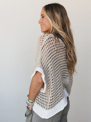 A side-back view of an open-knit crochet top in warm beige layered over a white shirt, styled with gray pants and stacked beaded bracelets — the kind of effortless, textured look that makes a Saturday feel intentional.