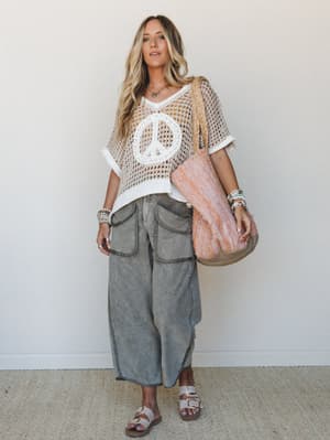 A breezy, full-body lifestyle shot of a model in a crochet peace-sign top layered over a white tank, paired with washed wide-leg cargo pants and sandals — the whole look reads effortless boho with a little vintage soul.