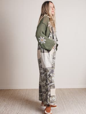 Woman wearing a green sweater with floral design and camouflage pants against a plain background