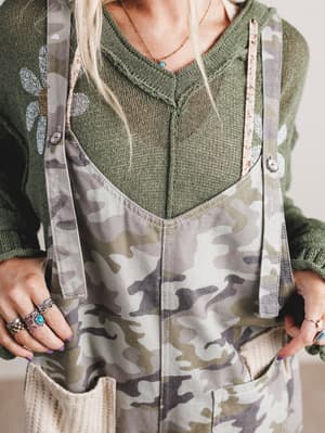 Person wearing a green sweater and camouflage overalls with a neutral background
