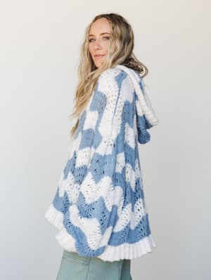 A breezy, oversized hooded cardigan in a wavy two-tone knit — cornflower blue and cream — styled over sage green pants, shot from behind in soft studio light with a quiet, free-spirited ease.