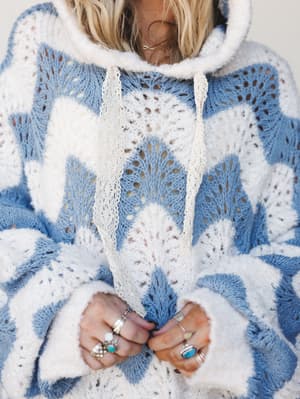 A close-up shot of a open-knit hooded sweater in cornflower blue and cream, styled with stacked silver and turquoise rings — the kind of piece that looks equally at home on a morning coffee run or a breezy beach walk.