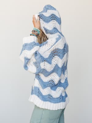 A side-back view of a slouchy open-knit hooded sweater in wavy cornflower blue and cream, styled with sage green pants and a stack of turquoise and beaded bracelets — the kind of sweater that makes a Tuesday feel intentional.